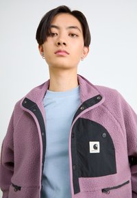 Joven con cabello corto y oscuro, vistiendo una chaqueta de forro polar morada con un bolsillo negro y una camisa de color azul claro, contra un fondo blanco.