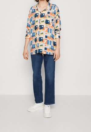 Färgglad skjorta med knappar och krage, med ett geometriskt mönster i blått, orange, grönt och beige; kombinerad med mörka jeans och vita sneakers.