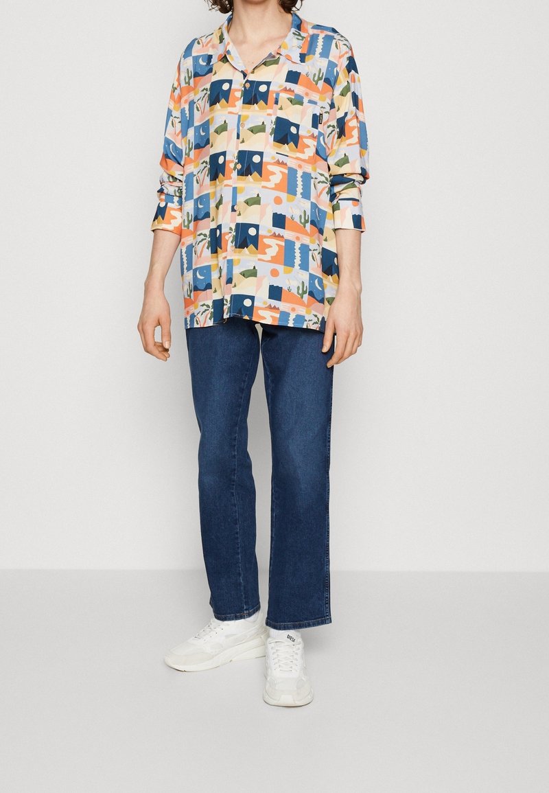 Chemise à boutons colorée avec col, présentant un motif géométrique en bleu, orange, vert et beige ; associée à un jean foncé et des baskets blanches.