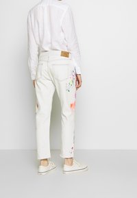 Bílé džíny s barevnými květinovými skvrnami od barvy. Mají zkrácený lem a koženou visačku na zadním pásku. Doplňují je bílé boty.