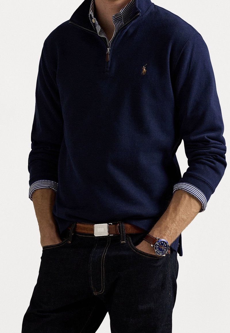 Homme portant un pull marine à fermeture éclair quart de longueur sur une chemise rayée, un jean foncé avec ceinture marron, et une montre marine avec bracelet en cuir marron, mains dans les poches.