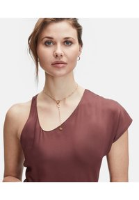 Bordeauxfarbene Bluse mit subtiler Sheen, ausgestattet mit einem runden Ausschnitt und kurzen Ärmeln, kombiniert mit einer zarten goldenen Kettenhalskette und einem runden Anhänger.