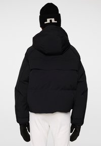 Svart pufferjacka med stor huva, med horisontella sömmar och rundad nederkant, tillsammans med en svart stickad mössa med vit detalj.