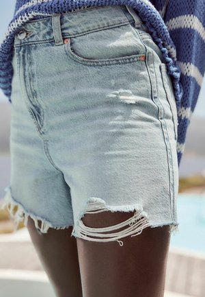 Shorts en denim bleu clair avec des bords effilochés et des détails usés sur les cuisses, portés avec un haut en tricot rayé bleu et blanc.