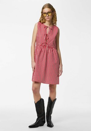 Jeune femme portant une robe rayée rouge et blanche sans manches avec des liens à l’avant, des lunettes teintées jaunes et des bottes de cowboy noires brodées.