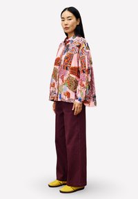 Frau steht da und trägt ein weites pinkes Shirt mit bunten abstrakten Mustern, burgunderfarbene Hosen und gelbe Schuhe vor einem weißen Hintergrund.