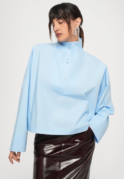 Light blue oversized shirt with a high collar, button front, and wide sleeves, paired with a glossy burgundy skirt.