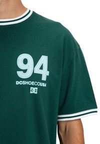 Zielona koszula z krótkim rękawem, posiadająca biały i ciemnozielony kołnierz oraz mankiety, z napisem "94 DCSHOECOUSA" i logo DC na klatce piersiowej.
