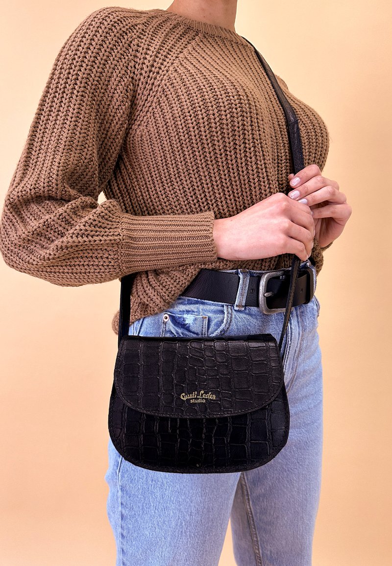 Osoba ubrana w brązowy, gruby sweter dziergany i jasnoniebieskie dżinsy, trzymająca czarną skórzaną torebkę na ramię z złotym napisem logo.