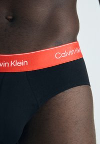 Schwarze Baumwollslips mit einem breiten roten Gummibund, auf dem "Calvin Klein" in Weiß gedruckt ist. Glatte Textur und körpernahe Passform.