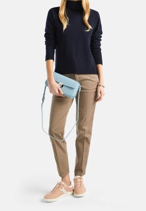 Donna che indossa un dolcevita blu navy, pantaloni khaki, sneakers rosa e tiene una borsa a tracolla azzurro chiaro per la tracolla davanti a uno sfondo bianco.