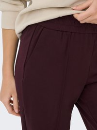 Gros plan d'une personne portant un pantalon à taille élastique violet foncé et un haut à manches longues beige, avec une main posée près de la poche.