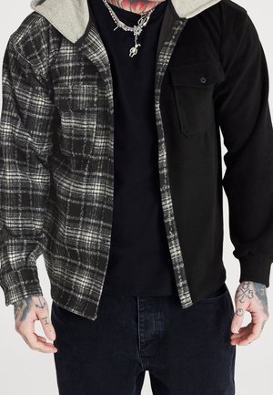 Mann trägt eine schwarze und graue karierte Kapuzenjacke mit einfarbig schwarzen Ärmeln und einer Fronttasche, kombiniert mit schwarzen Hosen und silbernen Kettenhalsketten.