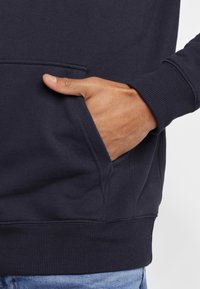 Dettaglio di una felpa blu navy che mostra una tasca laterale con la mano inserita, caratterizzata da polsini e vita a costine, realizzata in tessuto testurizzato.