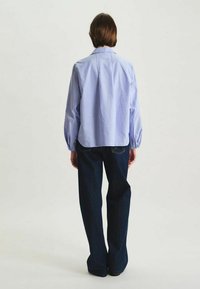 Chemise bleu clair à manches longues avec un pli dans le dos, assortie à un pantalon en denim foncé. La chemise possède un col classique et une fermeture à boutons.