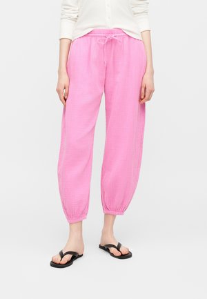 Osoba nosiaca voľné ružové nohavice so sťahovacou šnúrkou a elastickými manžetami, čierne žabky, stojaca na jednoduchom pozadí.