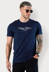 Navy blue fitted T-shirt in smooth fabric featuring white graphic text, crew neckline, short sleeves, and tattoo details on the arm.