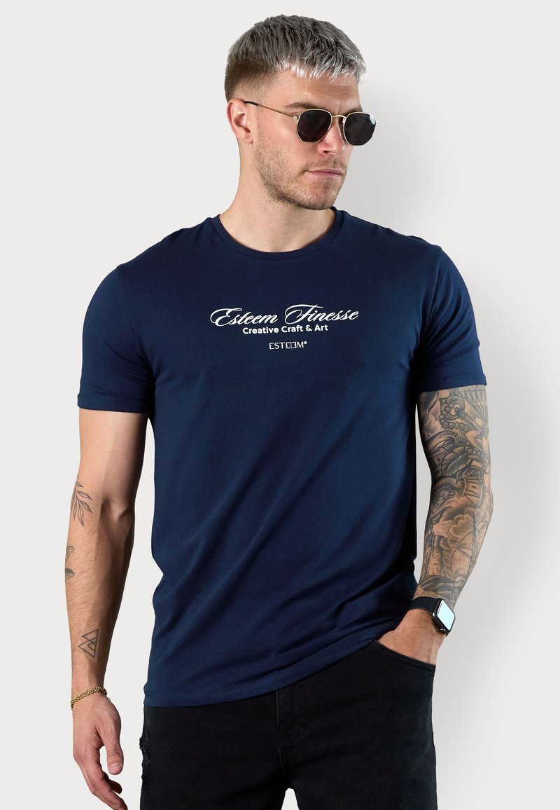 Navy blue fitted T-shirt in smooth fabric featuring white graphic text, crew neckline, short sleeves, and tattoo details on the arm.