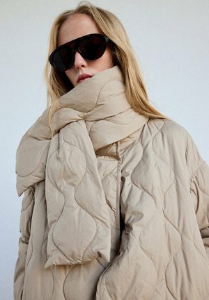 Blonde Frau, die eine übergroße beige gesteppte Jacke und einen passenden Schal mit schwarzen, übergroßen Sonnenbrillen vor einem einfarbigen hellen Hintergrund trägt.