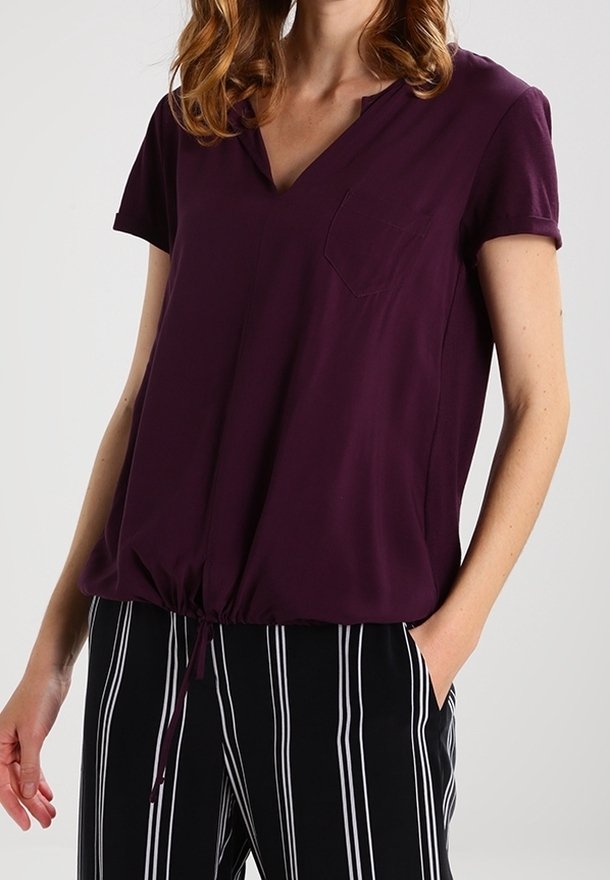 Femme portant un haut violet à manches courtes en V avec poche avant et un pantalon noir à rayures blanches verticales, une main dans la poche.