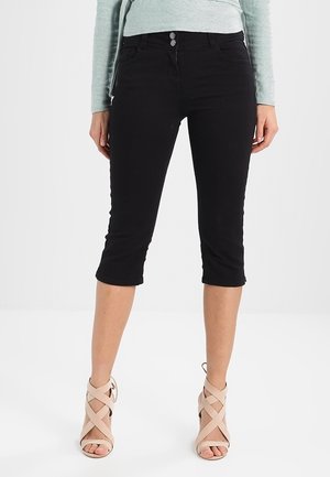 Femme portant un pantalon noir court avec double bouton et des sandales à talons hauts à lanières rose clair, debout sur fond blanc.