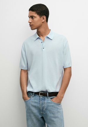 Ung mand iført en lyseblå kortærmet poloshirt og lyseblå jeans, hænderne i lommerne, kigger til sin venstre mod en ensfarvet baggrund.