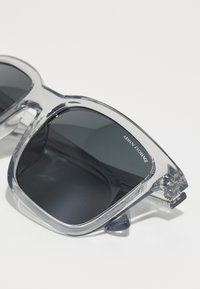 Ochelari de soare gri transparenți cu lentile întunecate, formă pătrată și brațe proeminente cu logo-ul "Armani Exchange." Textură netedă, detalii minime.