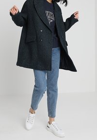 Abrigo largo de un verde oscuro y textura, con solapas anchas y botones dobles, llevado sobre una sudadera oscura, combinado con jeans de un azul claro y zapatillas blancas.