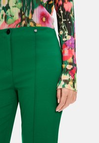 Grüne Hosen mit einer glatten Textur und Slim-Fit, ausgestattet mit einem Knopfverschluss. Kombiniert mit einem gemusterten, bunten Langarmoberteil.