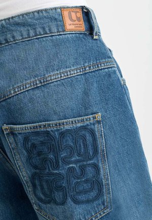 Denim jeans featuring a leather logo patch, a back pocket with embossed pattern, and a medium blue color with visible stitching details.