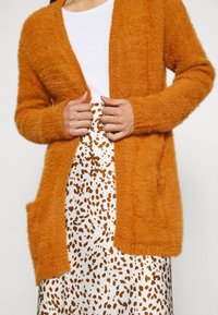 Pomarańczowy puszysty kardigan z długimi rękawami, z otwartym przodem i dwoma kieszeniami. Noszony na białym topie i białej spódnicy z brązowymi plamkami.