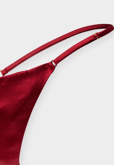 Ρυθμιζόμενο λεπτό λουράκι με χρυσή πόρπη που στερεώνεται σε ένα λείο ένδυμα από σκουρό κόκκινο σατέν ύφασμα, σε φόντο γκρι.