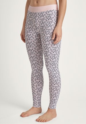 Vrouw in roze leggings met een klein marineblauw bladdessin en een brede roze tailleband, die blootsvoets staat op een effen achtergrond.