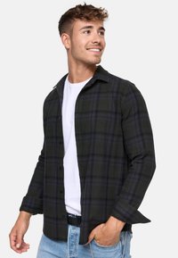 Camisa de franela en verde oscuro con patrón de cuadros en negro y púrpura, estilo de botones, mangas largas y una camiseta blanca debajo.