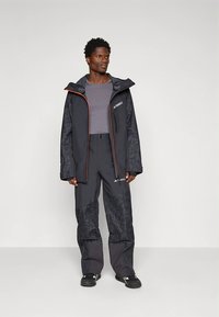 Vyriškas lauko drabužių komplektas, kuriame yra tamsiai pilka atspari vandeniui striukė su oranžiniais akcentais ir derančios kelnės su tekstūruotu raštu.