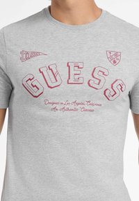 Hallide hallist puuvillane T-särgi, mille esiküljel on punane graafiline tekst "GUESS", asutamisaasta "1981" ja emblem "CLASSICS". Lühikesed varrukad, ümberlõigatud kaelus.