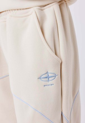 Beige joggers van zacht materiaal hebben een blauw geborduurd logo en contrasterende stiksels langs de naden, met zijzakken.