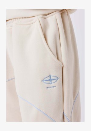 Joggers beige hechos de tejido suave presentan un logo bordado en azul y costuras en contraste a lo largo de las costuras, con bolsillos laterales.