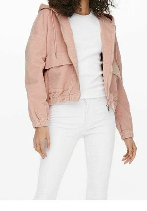 Vrouw draagt een lichtroze kort geribbelde jas over een wit T-shirt met witte broek, staat met ontspannen armen.