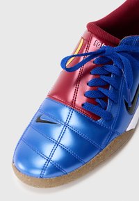 Sapatilha sintética azul e vermelha com textura acolchoada, logótipo da Nike em preto, atacadores azuis e sola de borracha castanha. Apresenta um design de cano baixo.