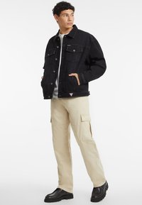 Giacca di jeans nera con due tasche frontali, cuciture chiare e un'etichetta a patch triangolare. Abbinata a pantaloni cargo beige e stivali neri.