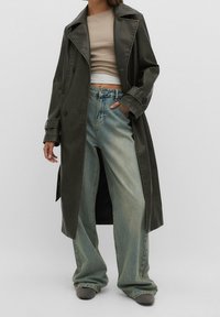 Długi, ciemnozielony płaszcz wykonany z gładkiego materiału, z dwoma dużymi guzikami, z wcięciem przy kołnierzu i rozcięciem po bokach. Noszony na szerokich, niebieskich dżinsach.