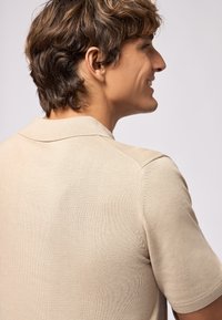 Polo shirt a maglia beige con una texture morbida, caratterizzato da un colletto classico e maniche corte, mostrato da un'angolazione laterale.