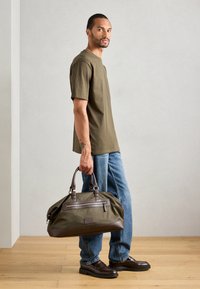 Bărbat îmbrăcat cu un tricou oliv, blugi albaștri și pantofi maro, ținând o geantă mare de voiaj oliv și maro în interior.