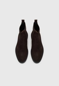 Botas de tobillo de ante marrón con una textura suave, paneles laterales elásticos y una suela de goma negra. Cuenta con una tira en la parte trasera para facilitar su colocación.