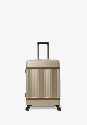 Zandkleurige harde koffer met verticale groeven, bruine afwerking, uitgeschoven zwarte telescopische hendel en vier zwenkwielen op een witte achtergrond.
