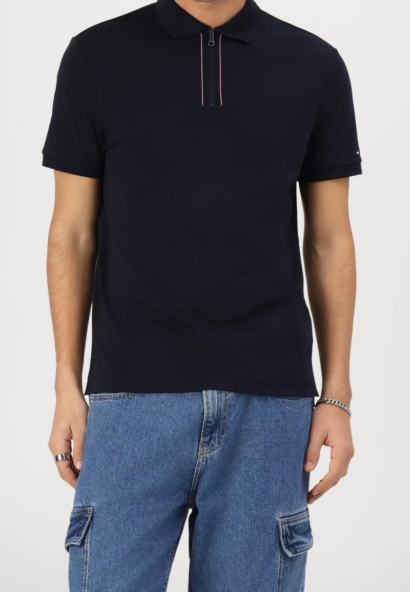 Homme portant un polo noir à manches courtes avec une fermeture éclair frontale et un pantalon cargo en jean bleu, sur un fond uni.