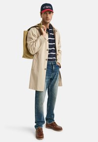 Trench coat beige, polo a righe blu navy, jeans blu, scarpe marroni e un cappellino blu navy con dettagli rossi. Borsa a tracolla appoggiata sulla spalla.