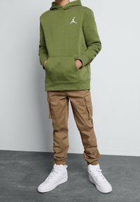 Felpa verde con cappuccio e tasca frontale, abbinata a pantaloni cargo beige con tasche laterali e sneakers bianche alte.