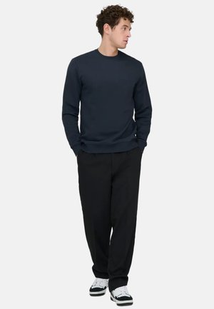 Mies, jolla on päällään laivastonsininen pusero, mustat housut ja valkoiset lenkkarit, seisoo taskut käsissä ja katsoo sivulle valkoisella taustalla.